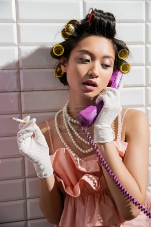 brune et asiatique jeune femme avec des bigoudis de cheveux debout en haut à volants rose, collier de perles et gants blancs fumer et parler sur le téléphone rétro près de tuiles blanches, femme au foyer, tenant cigarette 