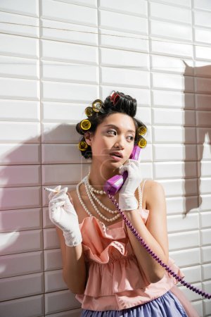 fashionable and asian young woman with hair curlers standing in pink ruffled top, pearl necklace and white gloves smoking and talking on retro phone near white tiles, housewife, holding cigarette 