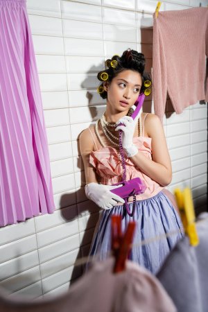 asian young woman with hair curlers standing in pink ruffled top, pearl necklace and white gloves, talking on purple retro phone and standing near clean laundry handing near white tiles, housewife 