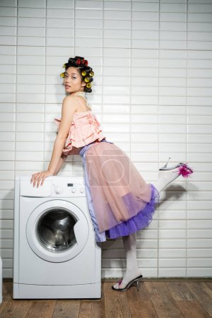 asiatische junge Frau mit Lockenwicklern in rosa Rüschenoberteil, Perlenkette, Tüllrock und Federabsätzen neben moderner Waschmaschine neben weißen Fliesen in der Waschküche, volle Länge 