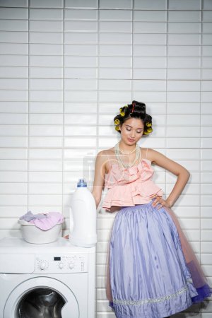 asiatische junge Frau mit Lockenwicklern, die mit der Hand auf der Hüfte in Rüschenoberteil steht, Perlenkette und Tüllrock in der Nähe von Waschschüssel mit schmutziger Kleidung auf moderner Waschmaschine mit Waschmittel in der Waschküche 