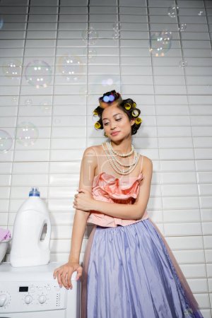 ładna azjatycka młoda kobieta z lokówkami stojącymi w falbanki góry, perłowy naszyjnik i spódnica tiulu w pobliżu nowoczesnej pralki z detergentem w pralni, gospodyni domowa, patrząc daleko, bańki mydlane