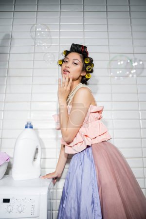 młoda Azjatka z lokówkami stojącymi w falbankach, perłowy naszyjnik i spódnica tiulu w pobliżu nowoczesnej pralki z butelką detergentu w pralni, gospodyni domowa, bańki mydlane
