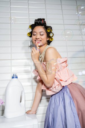 szczęśliwa i młoda Azjatka z lokówkami stojącymi w falbankach, perłowy naszyjnik i spódnica tiulu w pobliżu nowoczesnej pralki z butelką detergentu w pralni, gospodyni domowa, bańki mydlane