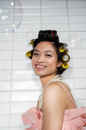 Porträt einer fröhlichen und jungen asiatischen Frau mit Lockenwicklern, die in Perlenkette neben verschwommenen Seifenblasen in der Waschküche mit weißen Fliesen steht, Hausfrau, natürliche Schönheit 
