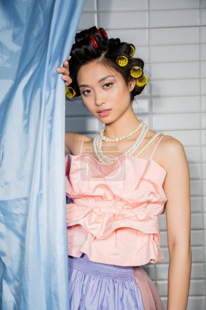 brunette and asian young woman with hair curlers standing in pink ruffled top with pearl necklace near blue bathroom curtain and looking at camera near white tiles at home 