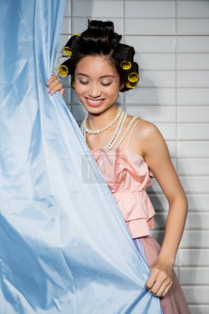 happy and asian young woman with hair curlers standing in pink ruffled top with pearl necklace near blue bathroom curtain and looking down near white tiles at home 