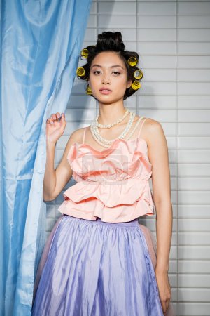 Foto de Mujer joven de moda y asiática con rizadores de pelo de pie en rosa con volantes superior con collar de perlas cerca de la cortina de baño azul y mirando a la cámara cerca de azulejos blancos en casa - Imagen libre de derechos