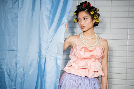 alluring and asian young woman with hair curlers standing in pink ruffled top with pearl necklace near blue bathroom curtain and looking away near white tiles at home 