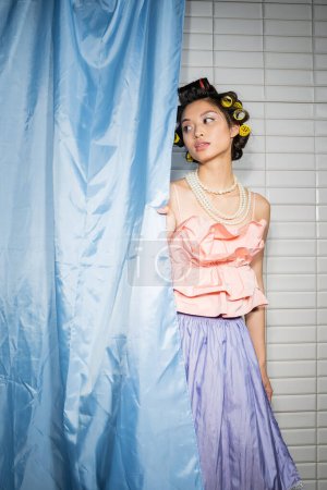 brünette und asiatische junge Frau mit Lockenwicklern, die in rosa Rüschentop mit Perlenkette und Rock neben blauem Badezimmervorhang steht und in der Nähe weißer Fliesen zu Hause wegschaut 