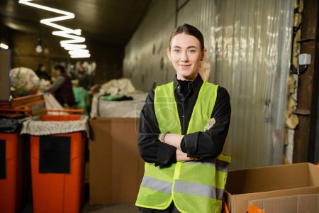 Young and smiling worker of waste disposal station in high visibility vest looking at camera and crossing arms while standing near blurred bins at background, garbage recycling concept