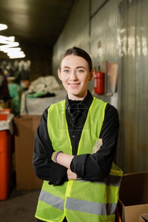Photo for Positive and young worker in gloves and high visibility jacket crossing arms and looking at camera while standing in blurred waste disposal station at background, garbage sorting and recycling concept - Royalty Free Image