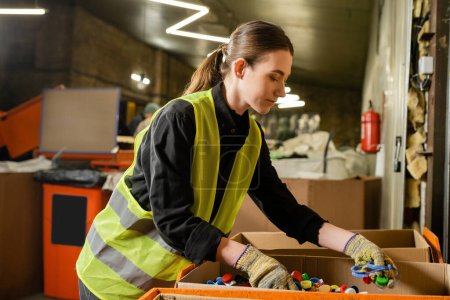 Photo for Young female worker of garbage sorting center wearing protective clothing and gloves while working with plastic caps in carton boxes, garbage sorting and recycling concept - Royalty Free Image