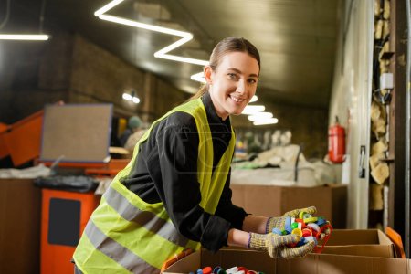 Clasificador femenino joven y sonriente en chaleco de alta visibilidad y guantes mirando a la cámara mientras sostiene tapas de plástico cerca de cajas de cartón y trabaja en el centro de clasificación de basura, concepto de clasificación de basura
