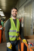 cheerful sorter in high visibility vest and protective gloves looking at camera while standing near plastic caps in carton boxes in waste disposal station, garbage sorting and recycling concept Stickers #658268952