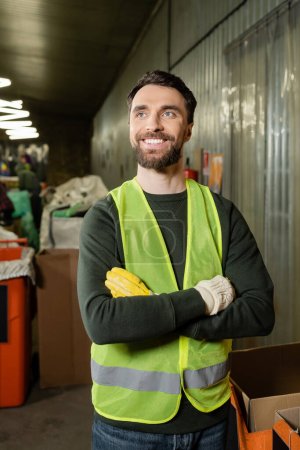Positiver männlicher Sortierer in Warnweste und Schutzhandschuhen, der die Arme verschränkt und im Stehen und bei der Arbeit im verschwommenen Müllentsorgungs- und Recyclingkonzept wegschaut
