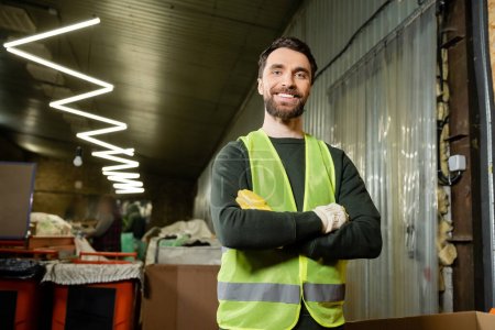 Photo for Cheerful worker in high visibility vest and protective gloves looking at camera and crossing arms while standing in blurred waste disposal station, garbage sorting and recycling concept - Royalty Free Image