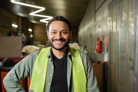 Photo for Portrait of cheerful indian sorter in high visibility vest looking at camera while standing and working in blurred waste disposal station, garbage sorting and recycling concept - Royalty Free Image