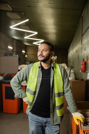 Photo for Indian man in high visibility vest and gloves looking away while standing near plastic caps in carton box and working in garbage sorting center, garbage sorting and recycling concept - Royalty Free Image