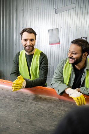 Fröhliche multiethnische männliche Sortierer in Schutzkleidung und Handschuhen stehen neben dem Fließband, während sie in Müllsortieranlagen, Müllsortier- und Recyclingkonzepten arbeiten
