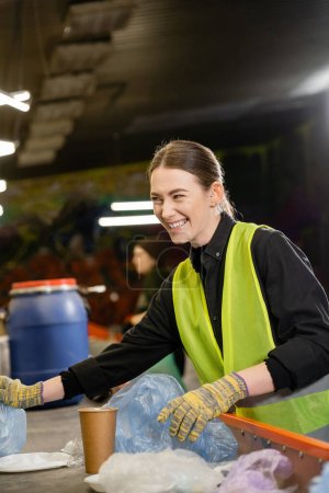 Cheerful young worker in protective vest and gloves sorting garbage on conveyor and looking away while working in waste disposal station, garbage sorting and recycling concept