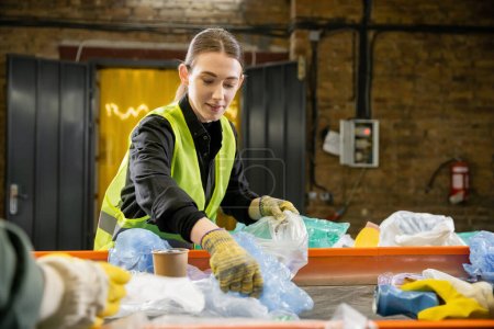 Joven trabajador sonriente en chaleco protector y guantes que clasifican la basura cerca del transportador mientras está parado en la estación de eliminación de residuos borrosos, clasificación de la basura y el concepto de reciclaje
