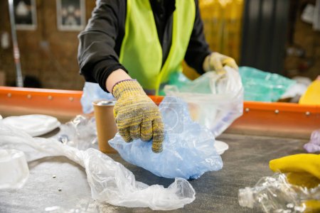 Verschwommene Ansicht eines Arbeiters in Schutzweste und Handschuhen, der Plastiktüten vom Band nimmt, während er im Hintergrund in der Müllentsorgungsstation arbeitet, Müllsortierkonzept 