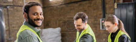 Positive indian worker in high visibility vest looking at camera while standing near blurred colleagues in garbage sorting center at background, garbage sorting and recycling concept, banner 