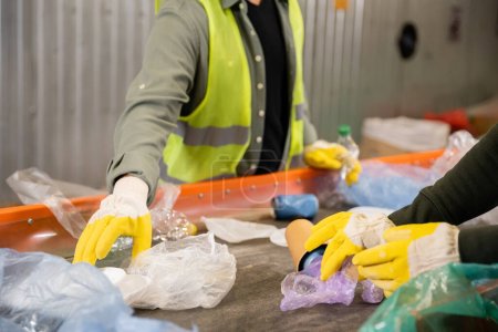 Vista recortada de los trabajadores con guantes protectores que toman basura plástica del transportador mientras trabajan juntos en un centro de clasificación de basura borrosa, clasificación de basura y concepto de reciclaje