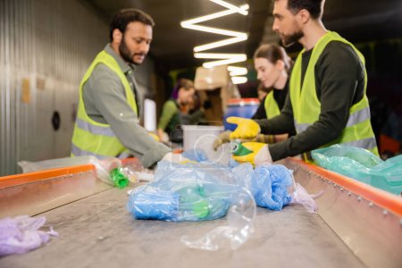 Plastikmüll auf Förderbändern in der Nähe verschwommener multiethnischer Arbeiter in Warnwesten und Handschuhen, die im Müllsortierzentrum, Müllsortier- und Recyclingkonzept zusammenarbeiten