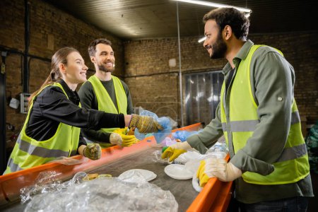 Smiling sorters in protective vests and gloves looking at indian colleague near plastic trash on conveyor while working in waste disposal station, garbage sorting and recycling concept