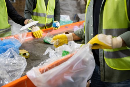 Cropped view of sorter in gloves and protective vest holding blurred plastic bag and taking trash from conveyor while working near colleagues in waste disposal station, garbage recycling concept