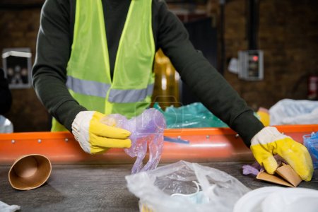 Cropped view of sorter in protective gloves and vest taking garbage from conveyor while working in blurred garbage sorting center, garbage sorting and recycling concept