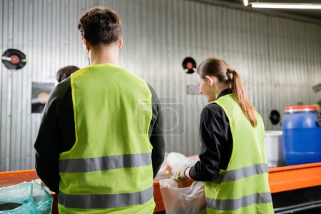 Worker in protective high visibility vest uniform separating trash near blurred colleagues and conveyor in garbage sorting center at background, garbage sorting and recycling concept