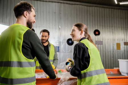 Smiling woman in protective uniform and gloves holding garbage and talking to interracial colleagues while working together near conveyor in waste disposal station, garbage sorting concept