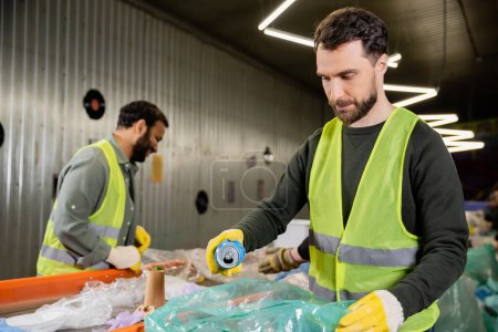worker in protective gloves and vest putting tin can in plastic bag while separating trash near multiethnic colleagues and conveyor in waste disposal station, garbage sorting and recycling concept