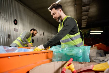 Bearded male sorter in protective vest and gloves taking garbage from conveyor near plastic bag while working near blurred indian colleague in waste disposal station, recycling concept