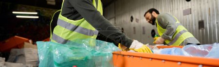 Male worker in protective gloves and reflective vest taking garbage from conveyor near plastic bag and blurred indian colleague at background in waste disposal station, recycling concept, banner 