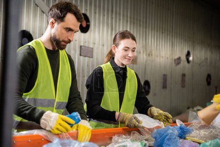 Lächelnder junger Arbeiter in Schutzhandschuhen und Sicherheitsweste trennt Müll auf Förderband, während er mit Kollegen gemeinsam in Entsorgungsstation arbeitet, Recyclingkonzept