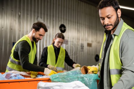 Bearded indian sorter in glove and high visibility vest putting paper cup in plastic bag while working near conveyor and blurred colleagues in waste disposal station, recycling concept
