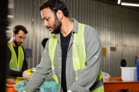 Bearded indian worker in high visibility vest separating trash while working near blurred conveyor and colleague in waste disposal station at background, recycling concept