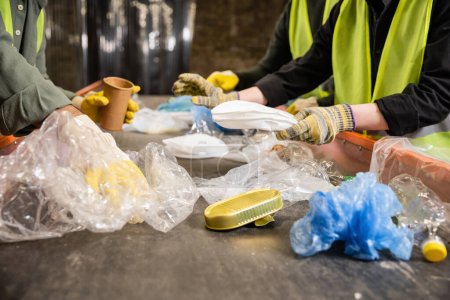Ausgeschnittene Ansicht von Arbeitern in Warnwesten und Handschuhen, die während ihrer Arbeit in der Entsorgungsstation verschiedene Plastik- und Papierabfälle auf dem Förderband trennen, Recyclingkonzept