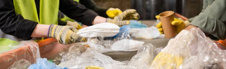 Cropped view of workers in protective gloves taking different plastic and paper trash from conveyor while working together in waste disposal station, recycling concept, banner 