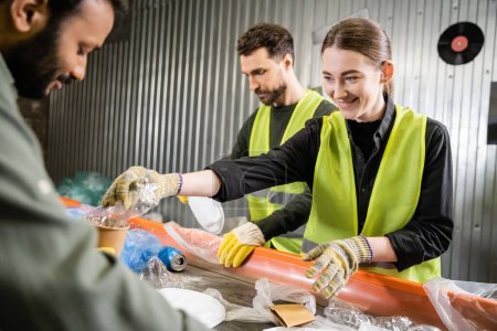 Positive worker in protective gloves and reflective vest taking plastic trash from conveyor near blurred interracial colleagues working in garbage sorting center, recycling concept