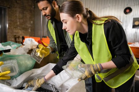 Young worker in protective gloves and safety vest holding plastic container near sack and indian colleague while separating trash in waste disposal station, garbage sorting and recycling concept