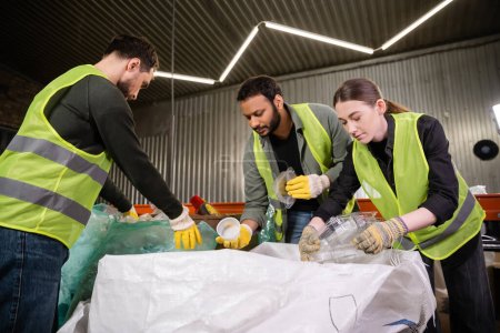 Young worker in reflective vest and gloves holding plastic containers near sack while sorting trash with multiethnic colleagues in waste disposal station, garbage sorting and recycling concept