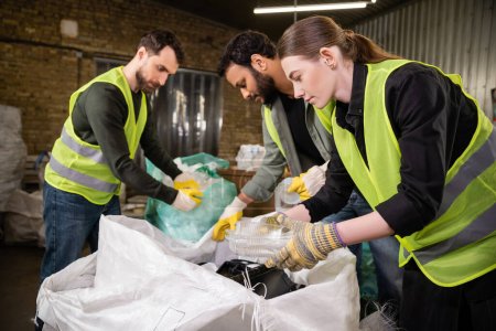 Young worker in safety vest and gloves separating plastic trash for recycling near sack and blurred interracial colleagues working in waste disposal station, garbage sorting and recycling concept