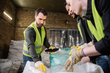 Bearded worker in high visibility vest and gloves taking garbage from sack for recycle while working with interracial colleagues together in waste disposal station, recycling concept