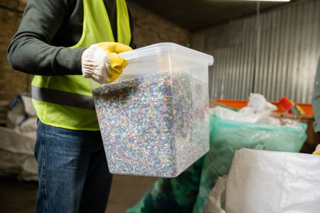 Ausgeschnittene Ansicht des Arbeiters in Warnweste und Handschuhaufbewahrungsbehälter mit Kunststoff zum Recyceln während der Arbeit im Müllsortierzentrum, Müllsortier- und Recyclingkonzept