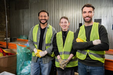 Positive Arbeiter in Warnwesten und Handschuhen, die in die Kamera schauen, während sie in der Nähe des Förderers stehen und in der Entsorgungsstation arbeiten, Recyclingkonzept